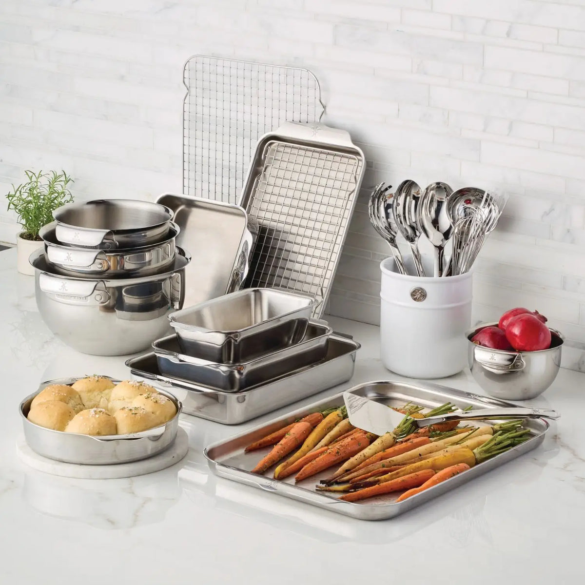 A kitchen scene showcases Hestan Provisions OvenBond Tri-ply Epicurean Stainless Steel 21-Piece Set, with mixing bowls, baking sheets, and a cooling rack. Durable bakeware holds roasted carrots and bread rolls. Utensils in a holder and bowls of radishes complete this culinary setup.