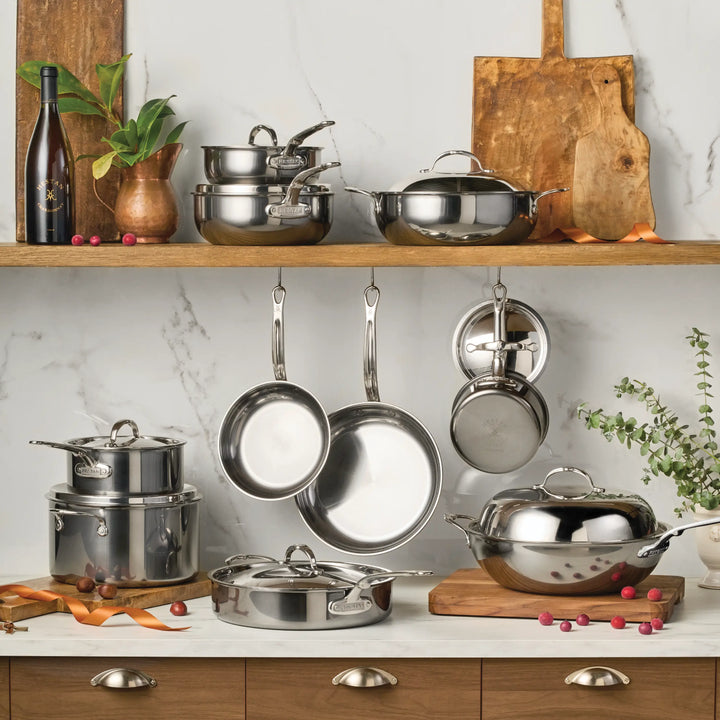 A kitchen displays the NanoBond 18-Piece Limited Edition Titanium Cookware Set with Chef's Pan—pots, pans, and kitchenware arranged on wood shelves and countertops with cutting boards, a plant, and olive oil against a marble backsplash.