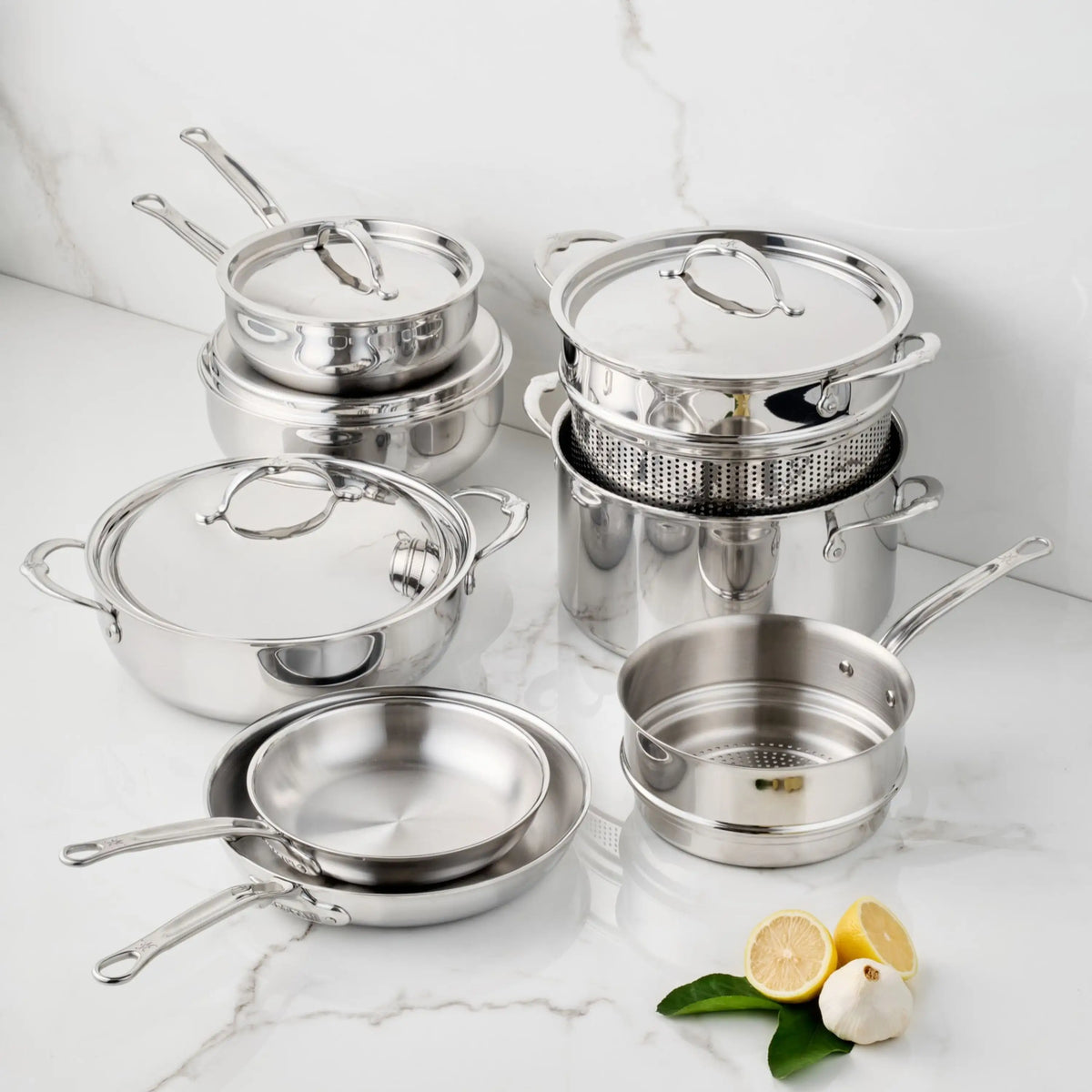 A 12-piece ProBond Luxe® Polished Clad Stainless Steel Set of pots, pans, lids, and a steamer insert arranged on a marble surface with garlic, lemon halves, and green leaves.