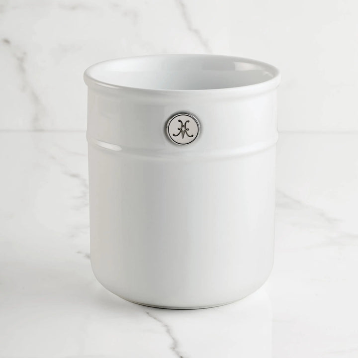 The Hestan Provisions Porcelain Bain Marie, with a round emblem bearing a stylized K, sits on white marble—ideal for stylishly organizing chef’s tools.