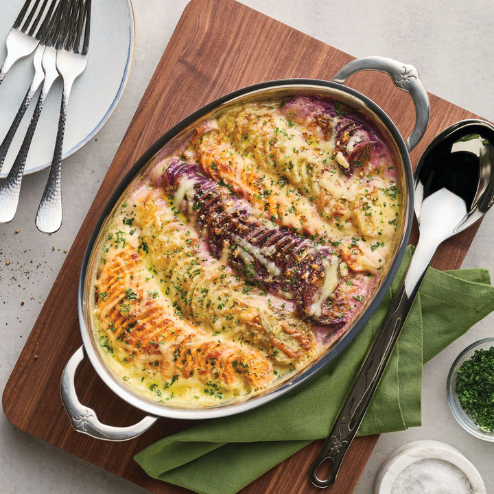 A 15-Inch x 9-Inch Oval Au Gratin baker by Hestan Provisions holds a cheesy vegetable casserole and rests on a wooden board, paired with a large serving spoon and utensils.
