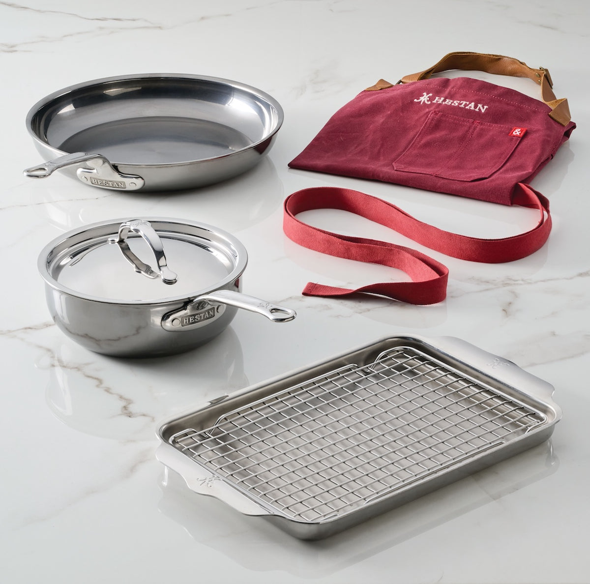 The NanoBond Titanium Grilling Essentials Cookware Set, 6-piece, includes a frying pan, lidded pot, roasting tray with rack, and a maroon grilling apron with tan straps—all displayed on white marble.