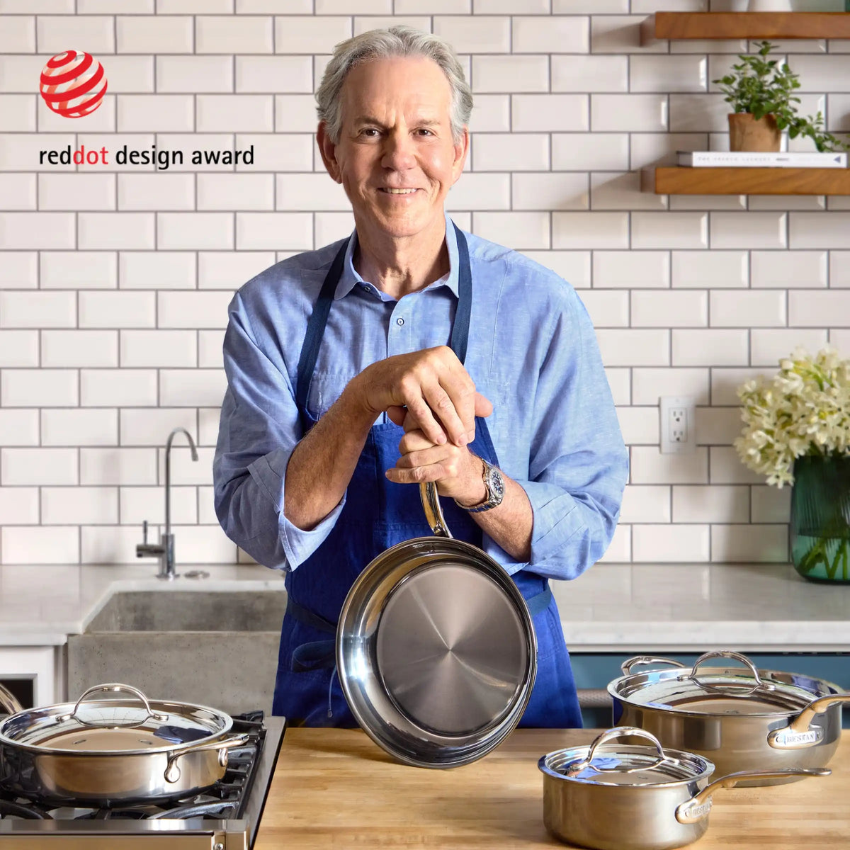 A man in a blue apron stands in a modern kitchen, holding a NanoBond Titanium Skillet. Several pots with lids are on the counter, and the Red Dot Design Award logo appears on the tiled wall in the background.