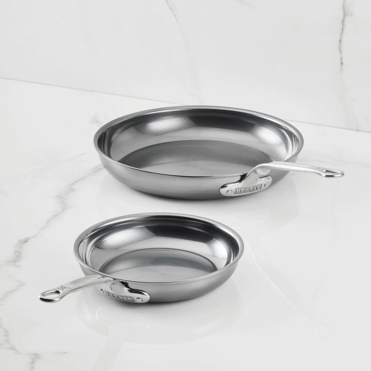 Two NanoBond Titanium Skillets with handles sit on a white marble surface. The different-sized pans feature a polished, reflective finish—ideal non-toxic cookware inspired by the Hestan NanoBond skillet.