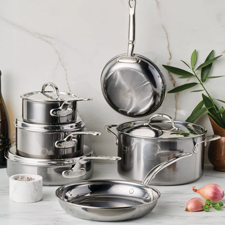 The NanoBond Titanium x Tyler Florence 41-Piece Cookware Set is displayed on a marble kitchen counter. Included are pots and pans, some with lids, alongside two shallots, a bowl of salt, an olive oil bottle, and a potted plant for an elegant touch.
