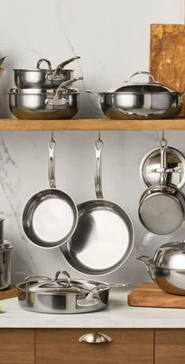 A kitchen features the NanoBond 18-Piece Limited Edition Titanium Cookware Set with Chef's Pan on a wooden shelf and counter, with pots, pans, lids, cutting boards, a bottle, and fresh herbs accenting a marble wall backdrop.