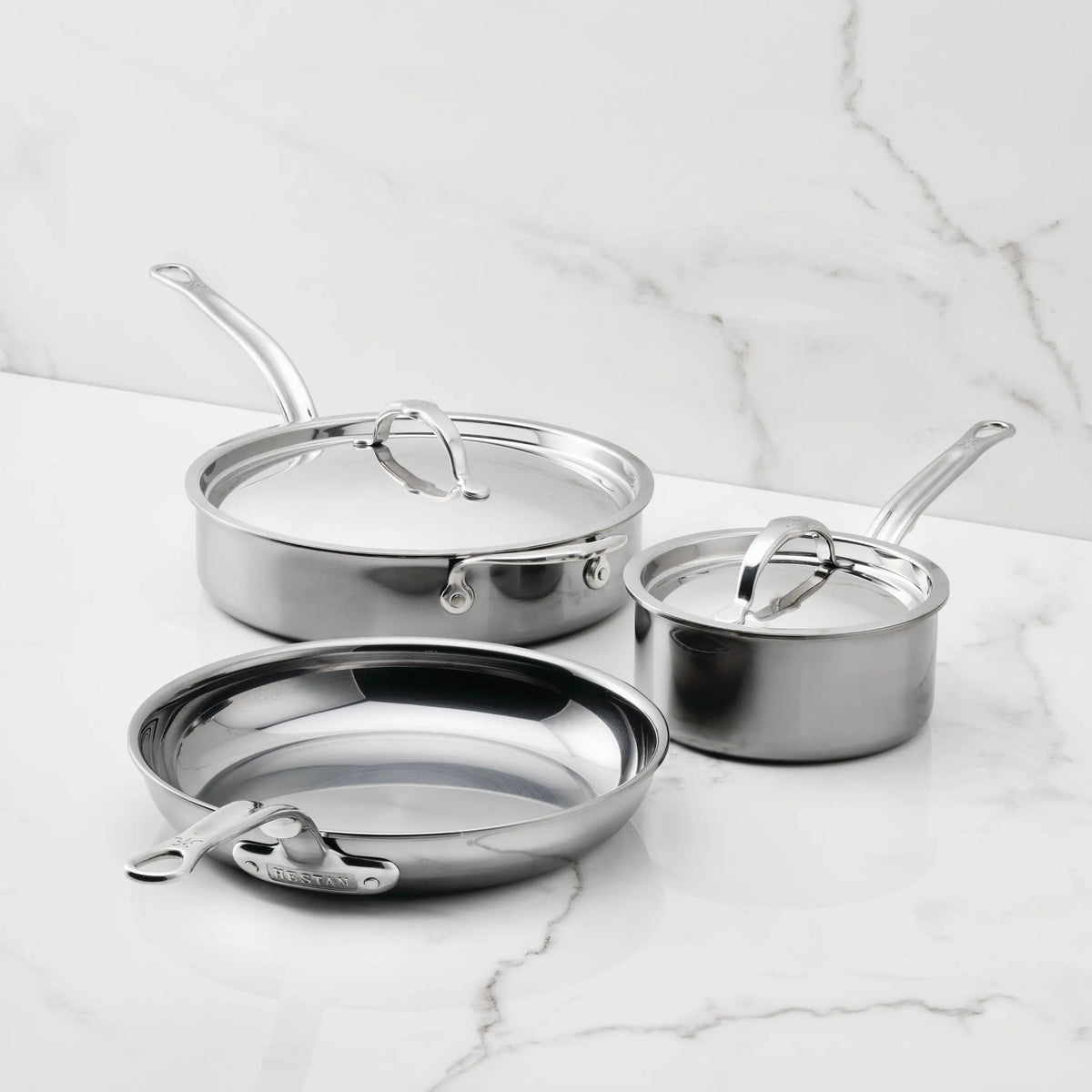 Showcased on a white marble surface, the NanoBond 5-Piece Titanium Essential Cookware Set features a sauté pan with lid, a saucepan with lid, and a frying pan—all designed with curved handles and polished finishes. Each piece embodies the advanced innovation of NanoBond Molecular Titanium for outstanding durability and performance.