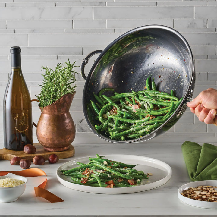 Someone pours green beans with cheese and bacon from a NanoBond Titanium Chef's Pan, 14-Inch, onto a white plate. Visible are a wine bottle, copper pitcher, rosemary, chestnuts, sliced almonds, and a green napkin.