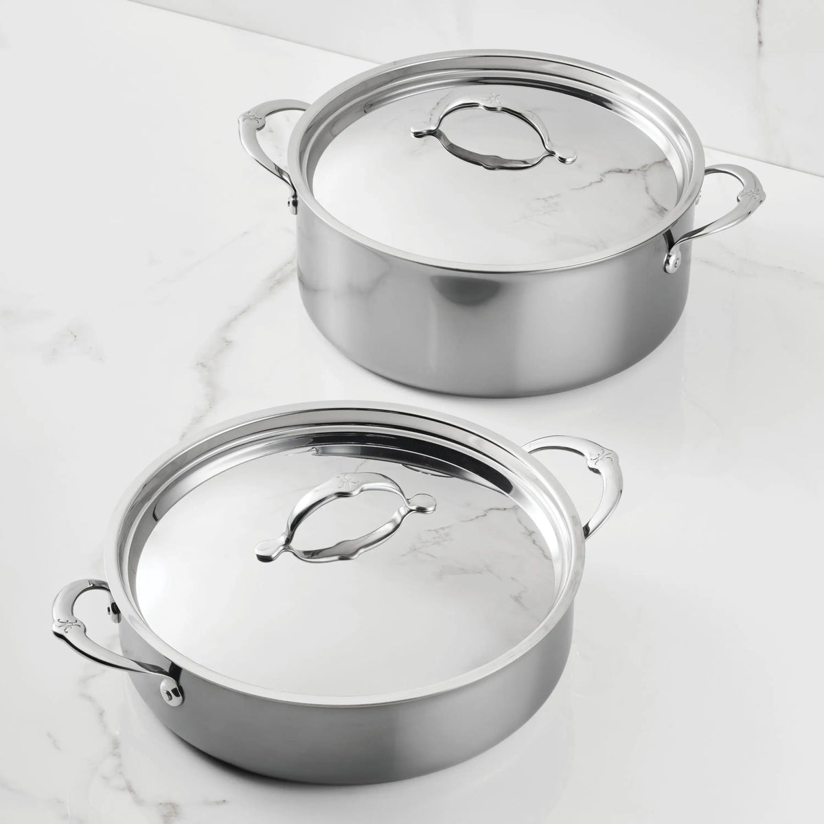 Two NanoBond Titanium Rondeaus with lids and handles are set on a white marble surface. The reflective cookware is positioned slightly apart, one rondeau in the foreground and one in the background.
