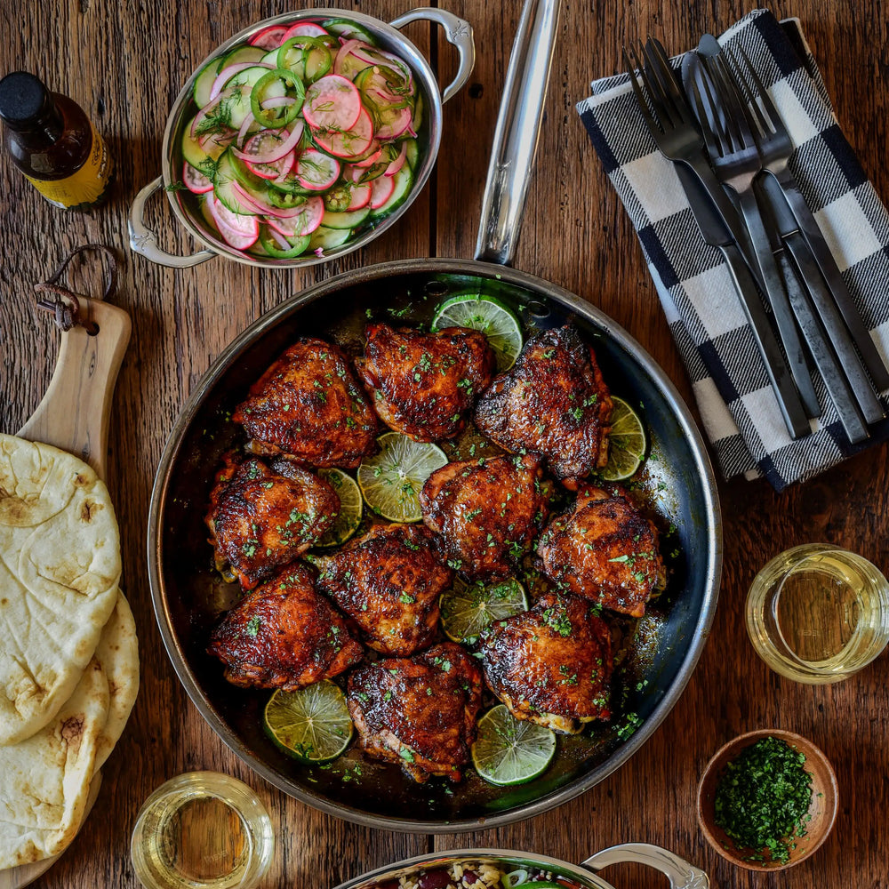 A NanoBond Titanium Skillet with cooked chicken thighs on lime slices sits on a wooden table, surrounded by cucumber and radish salad, stacked flatbreads, utensils on a napkin, and two glasses of white wine.
