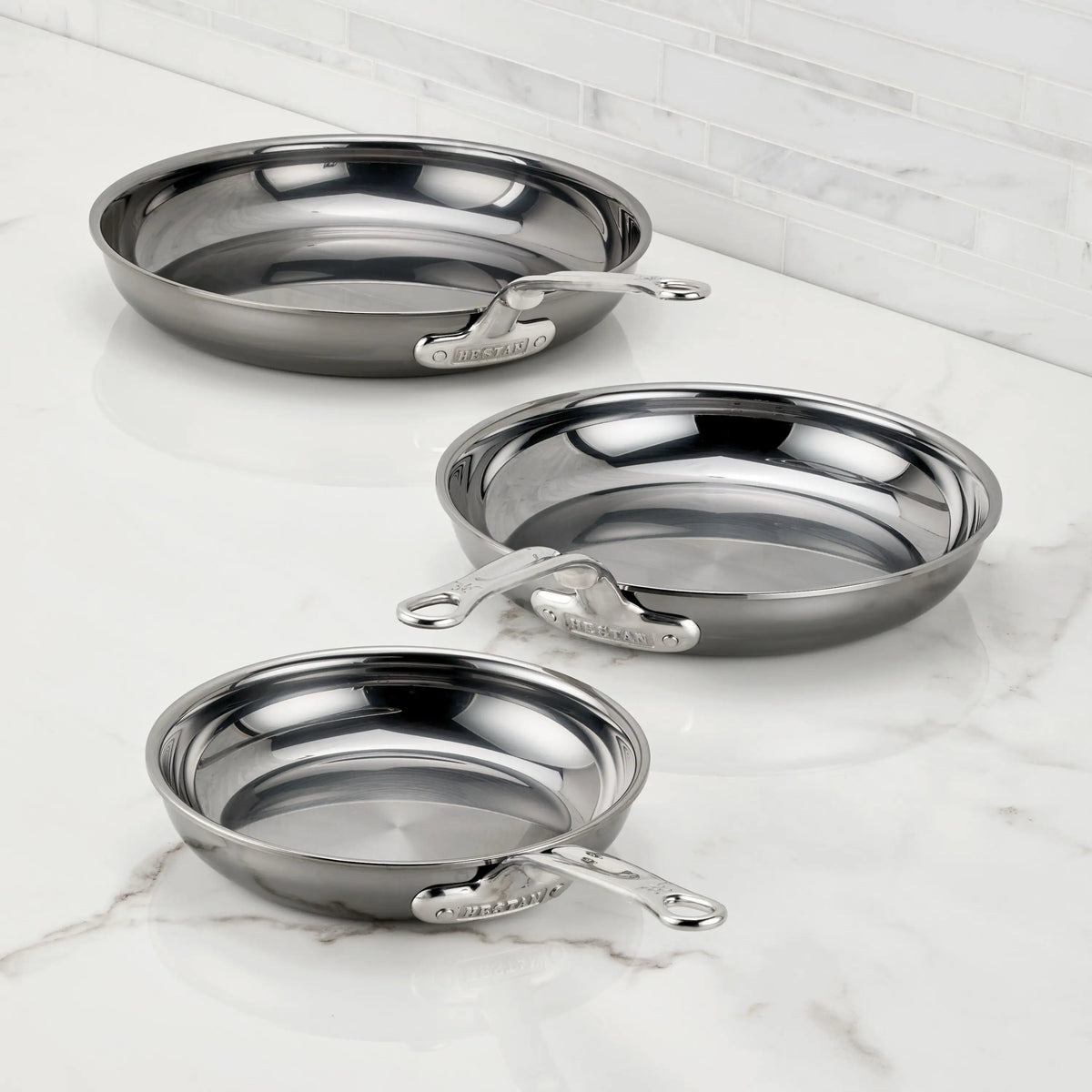 Three NanoBond Titanium Skillets in various sizes are displayed on a white marble countertop with a white tile backsplash, showcasing their shiny exteriors and sleek metal handles.