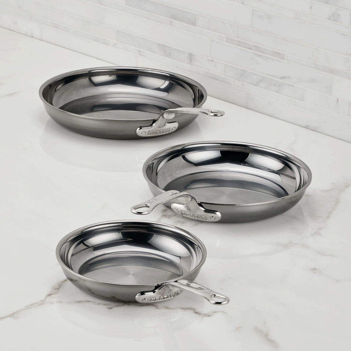 Three NanoBond Titanium Skillets in various sizes are displayed on a white marble countertop with a tiled backsplash. Each frying pan showcases a sleek metallic handle and non-reactive titanium construction.