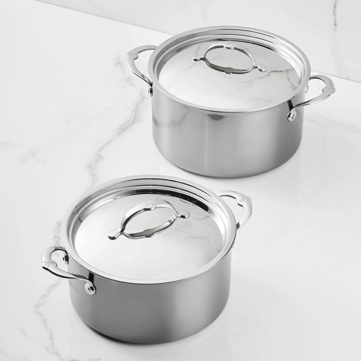 Two Titanium Stockpots from NanoBond, each with two side handles and topped with matching lids that have large looped handles, are displayed on a white marble surface.