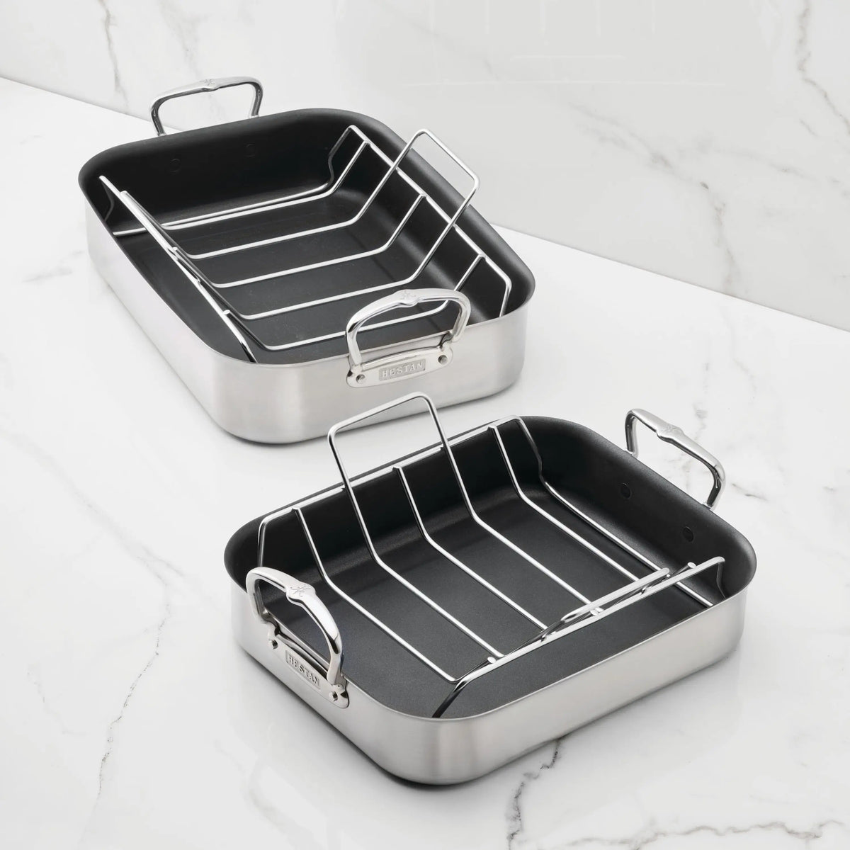 Two Hestan Provisions Nonstick Clad Stainless Steel Roasters with racks sit on a white marble countertop. These durable roasters feature sturdy handles and a black nonstick interior for effortless roasting and cleanup.