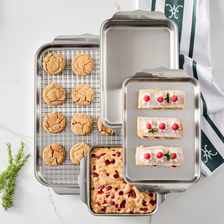 The OvenBond Tri-ply 5-Piece Set includes a cooling rack with cookies, a rectangular cake pan with a berry dessert, an empty rectangular pan, and a smaller tray with three rectangular pastries.