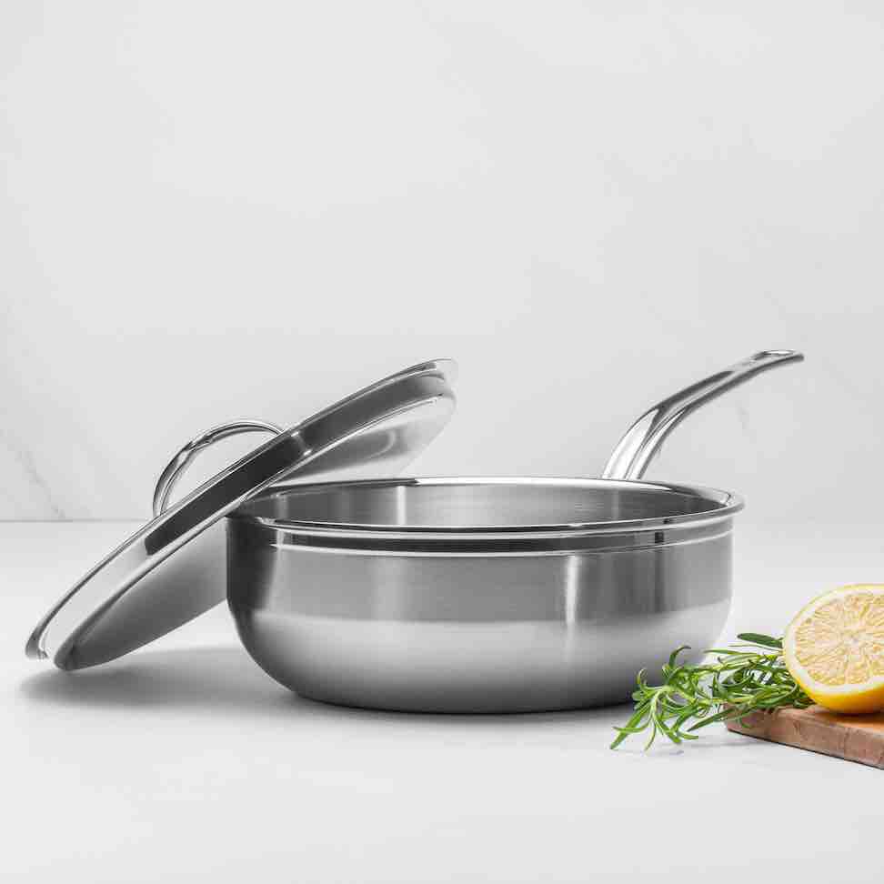 A saucepan from the ProBond Professional Clad Stainless Steel Induction 4pc Cookware Set, with a matching lid and right-extending handle, is placed on a white surface. A halved lemon and rosemary sprigs sit on a wooden cutting board nearby, enhancing its elegant look.