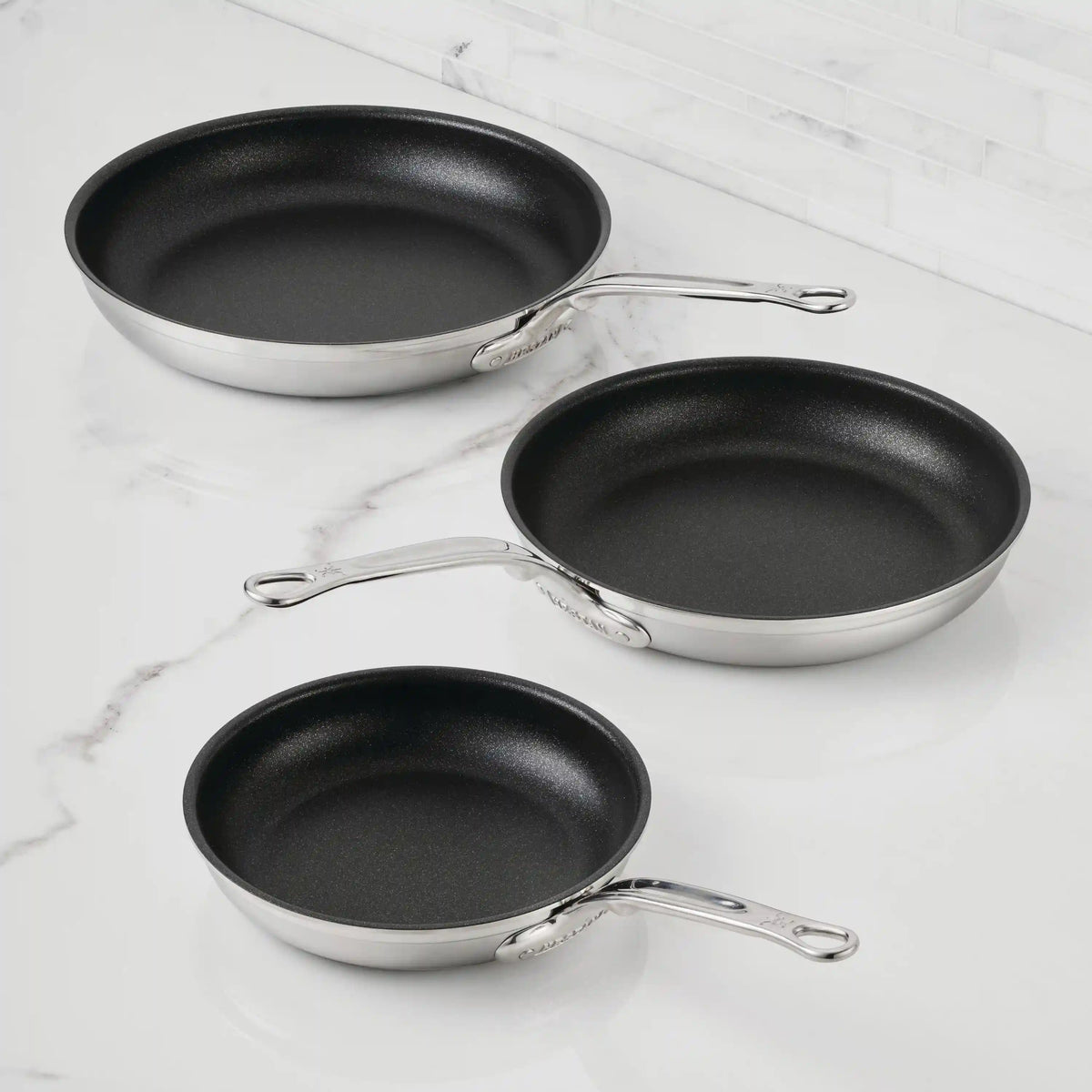 Three ProBond® Forged™ Professional Clad Stainless Steel TITUM® Nonstick Skillets in different sizes rest on a white marble surface, handles to the right. The TITUM® Nonstick System ensures easy cooking, set against a white tile wall.