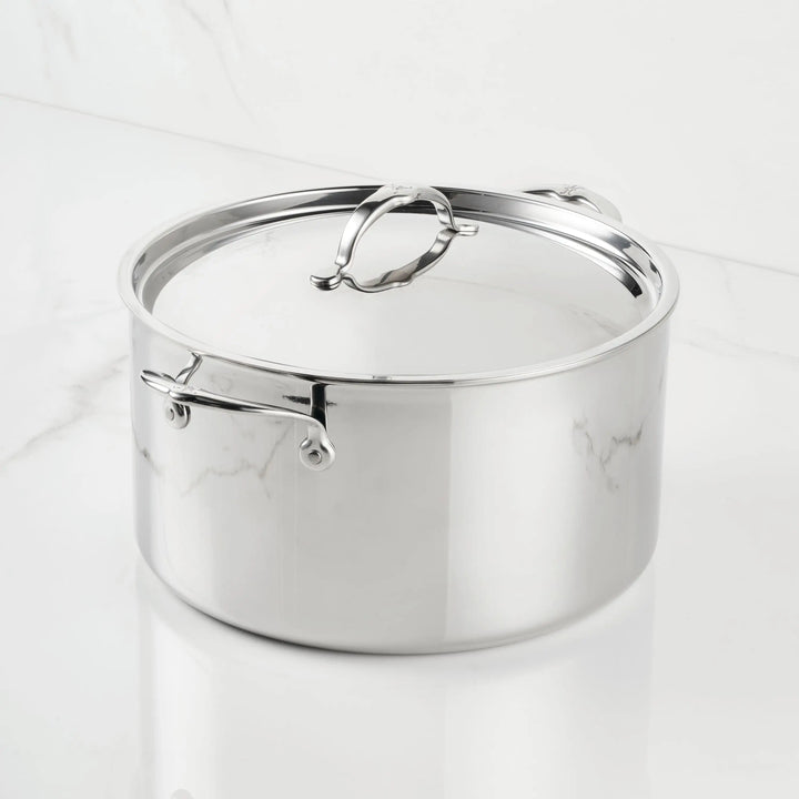 A ProBond® Luxe™ Polished Clad Stainless Steel Stockpot with lid, highlighting fine Italian craftsmanship and metal handles, is displayed on a white marble surface.