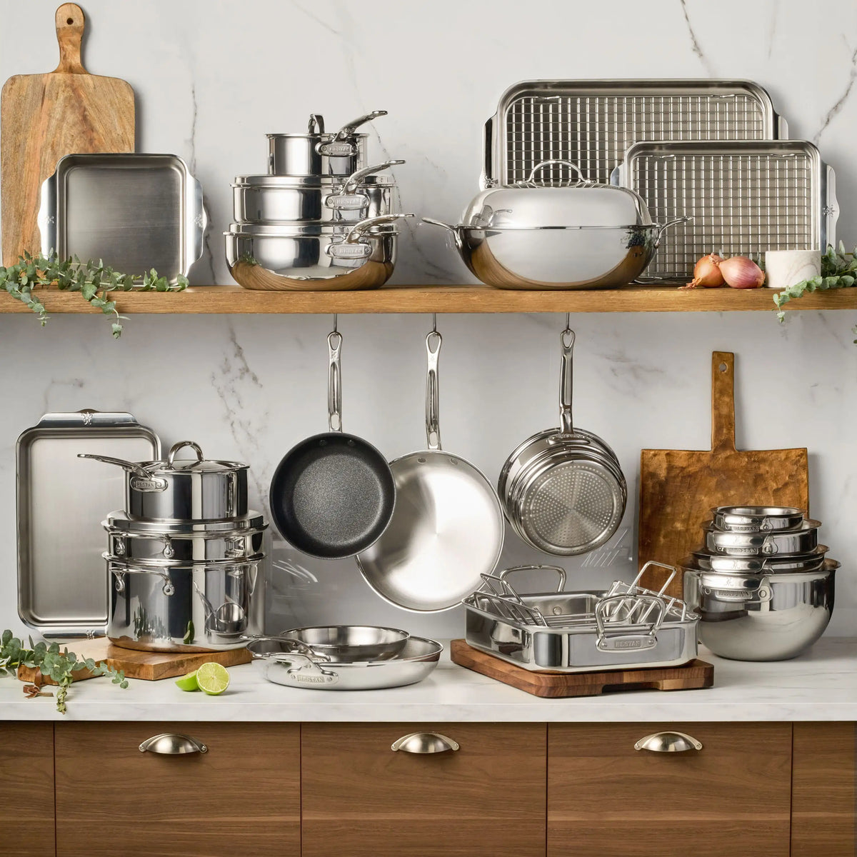 The ProBond® Luxe™ Polished Clad Stainless Steel Pristine Set, 29-Piece, is arranged on shelves and hooks with cutting boards, a lime, and vegetables, all set against a marble backsplash.