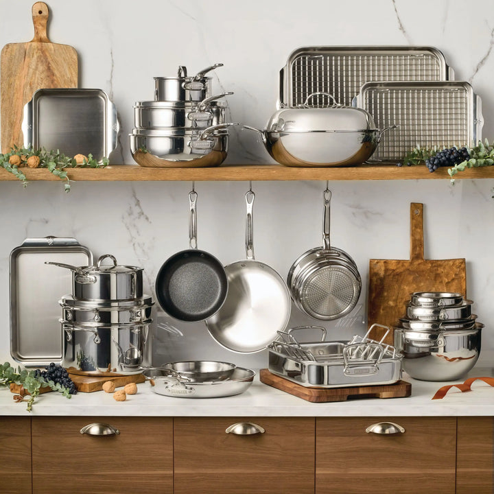 A kitchen scene features wood cabinets with silver handles and shelves displaying the ProBond® Luxe™ Polished Clad Stainless Steel Pristine Set, 29-Piece, including tri-ply pots, pans, bowls, and trays. Cutting boards, herbs, and grapes add decorative touches.