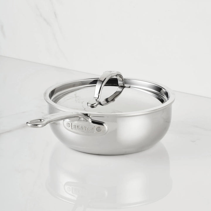 The ProBond® Luxe™ Polished Clad Stainless Steel Saucier Pan, 2-Quart, with lid and sturdy handle, sits on a white marble countertop against a matching marble wall.