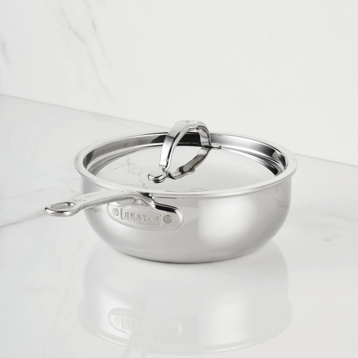 The ProBond® Luxe™ Polished Clad Stainless Steel Saucier Pan, 2-Quart, with lid and sturdy handle, sits on a white marble countertop against a matching marble wall.