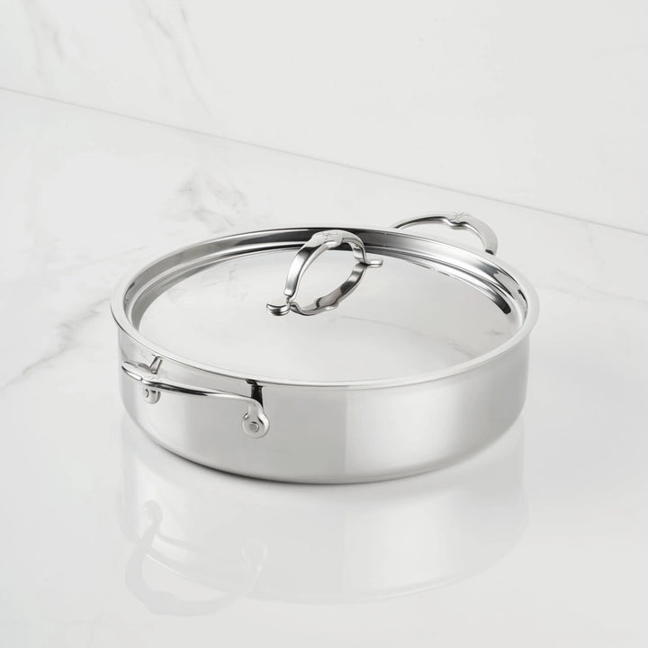 A ProBond® Luxe™ Polished Clad Stainless Steel Rondeau with two side handles and a matching lid is shown on a white marble surface, highlighting its professional design and easy-to-use features.