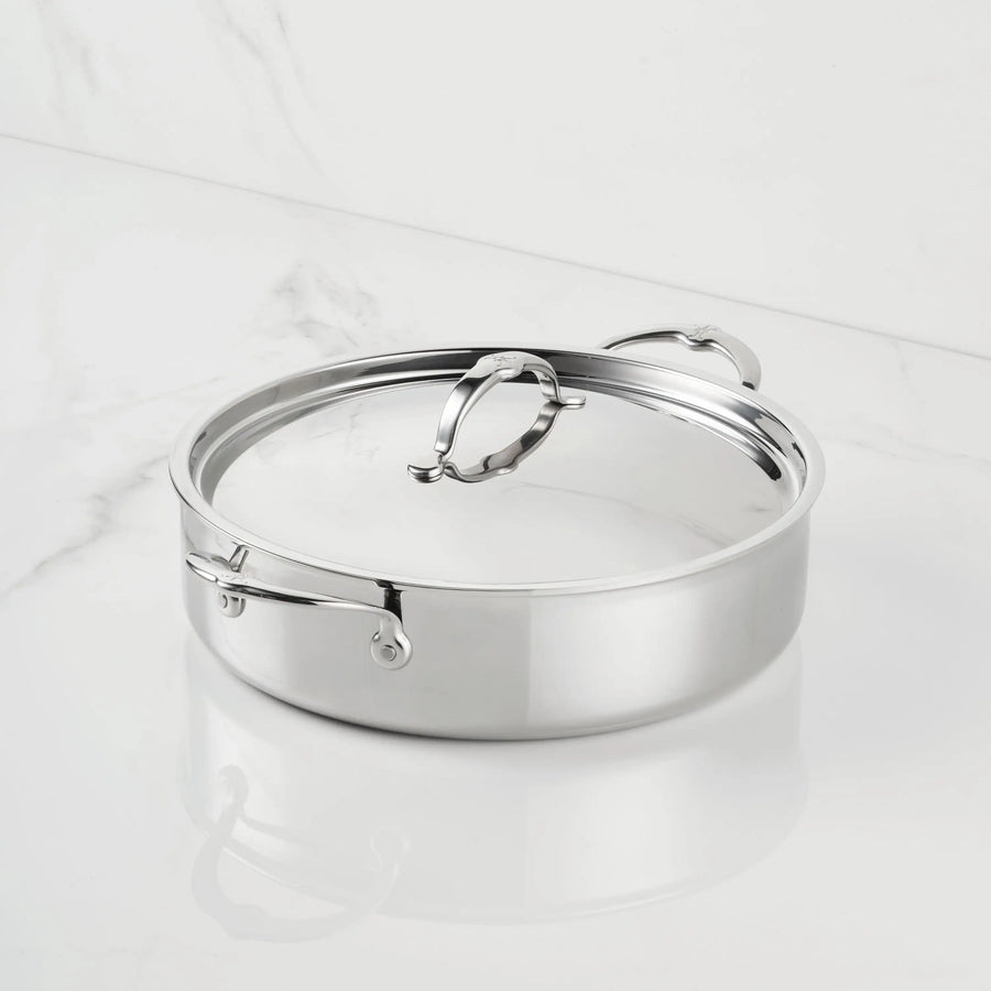 A ProBond® Luxe™ Polished Clad Stainless Steel Rondeau with two side handles and a matching lid is shown on a white marble surface, highlighting its professional design and easy-to-use features.