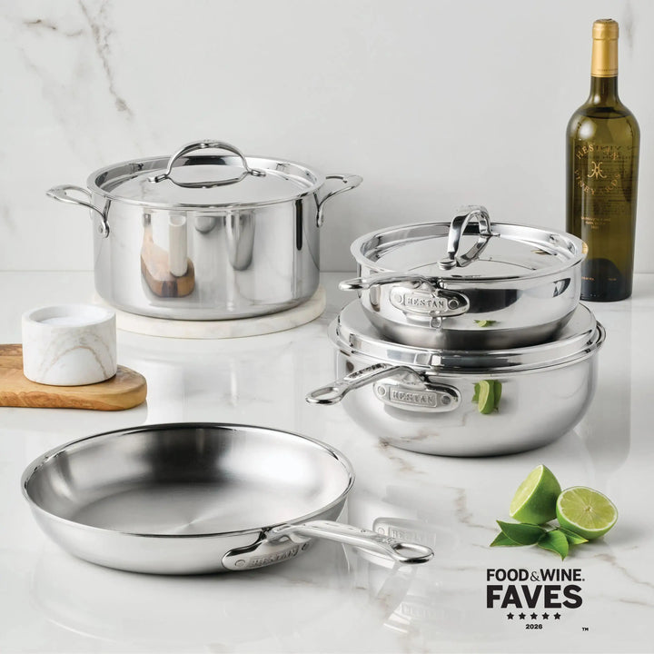 A collection of ProBond Luxe Polished Clad Stainless Steel Rondeaus with Lid, including a large lidded pot, two stacked lidded pots, and a shallow skillet, arranged on a marble surface with a bottle of wine and lime slices.