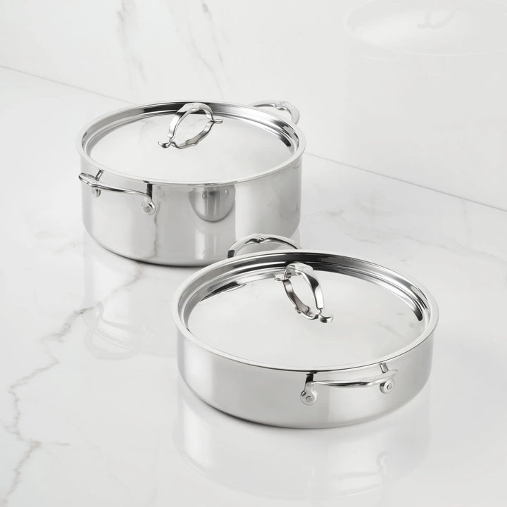 Two ProBond® Luxe™ Polished Clad Stainless Steel Rondeaus with matching lids sit on a white marble countertop, their reflective surfaces and sturdy handles enhancing the clean, minimalistic setting.