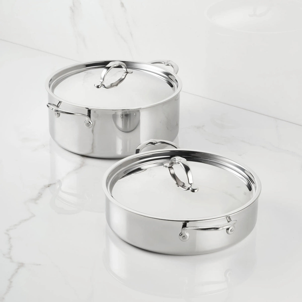 Two ProBond® Luxe™ Polished Clad Stainless Steel Rondeaus with matching lids sit on a white marble countertop, their reflective surfaces and sturdy handles enhancing the clean, minimalistic setting.