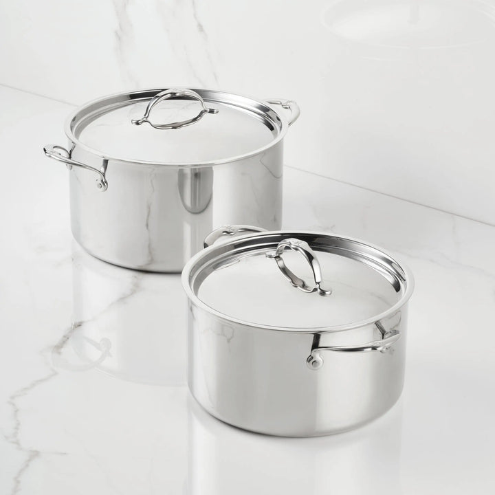 Two ProBond® Luxe™ Polished Clad Stainless Steel Stockpots with lids are placed side by side on a white marble countertop, their reflective finish adding a sleek, professional touch to the kitchen.