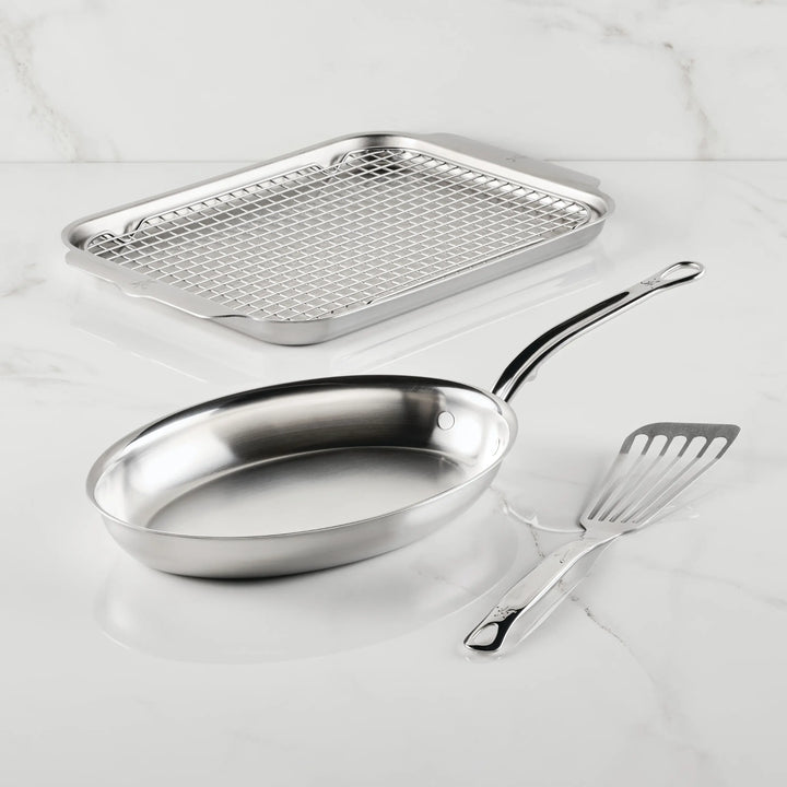The Hestan Culinary Provisions Fish Pan Set, 4-Piece, featuring a stainless steel frying pan, metal slotted spatula, rectangular baking tray, and wire cooling rack, is displayed on a white marble surface.