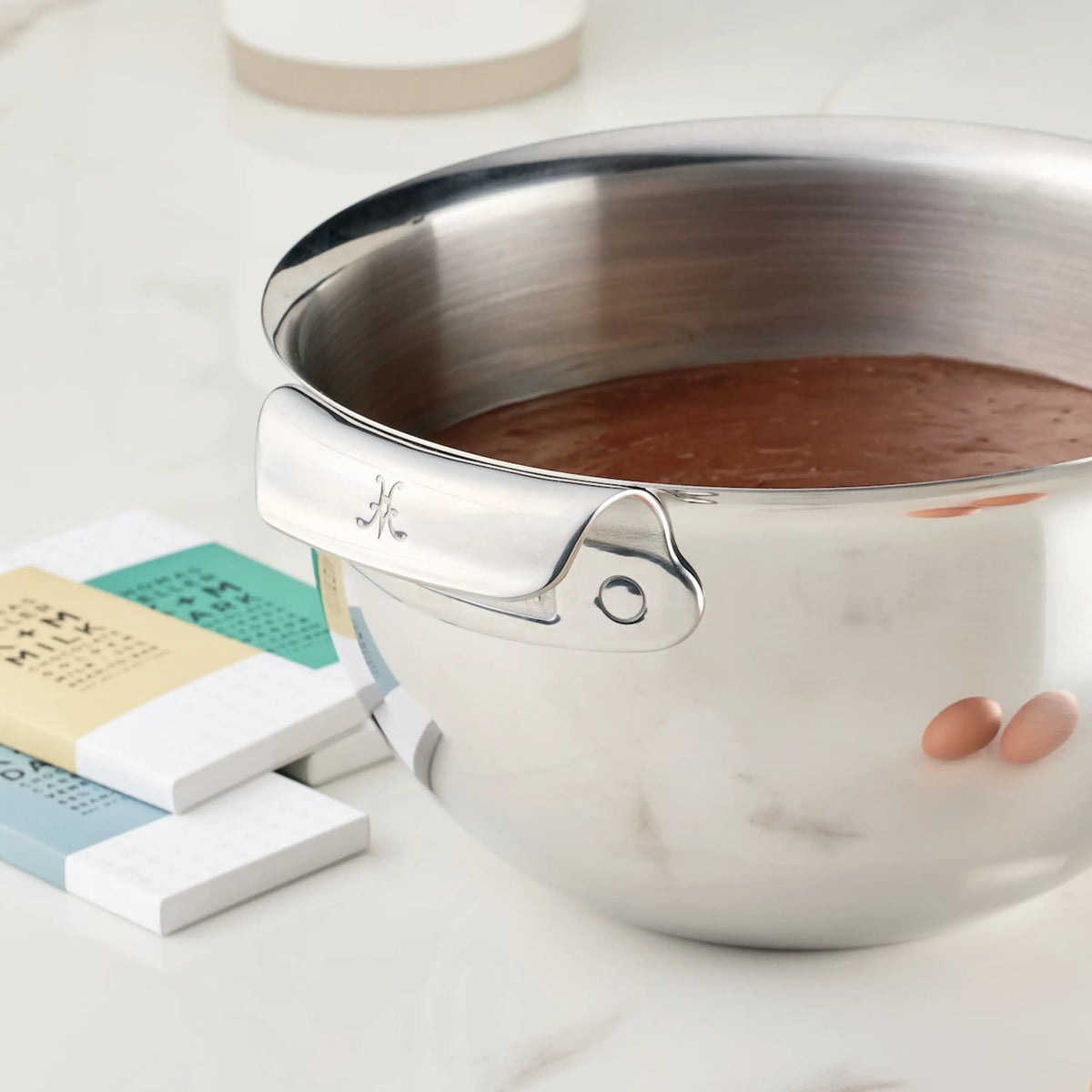 A Hestan Provisions Stainless Steel Mixing Bowl, 7-Quart, filled with chocolate batter sits on a white countertop. Three chocolate bars in pastel wrappers are placed beside the bowl.