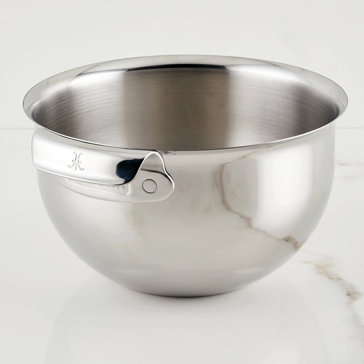A Hestan Provisions 7-Quart Stainless Steel Mixing Bowl with a rolled rim and side handle sits on white marble. Dishwasher safe, it has a reflective finish and an engraved letter K on the handle.