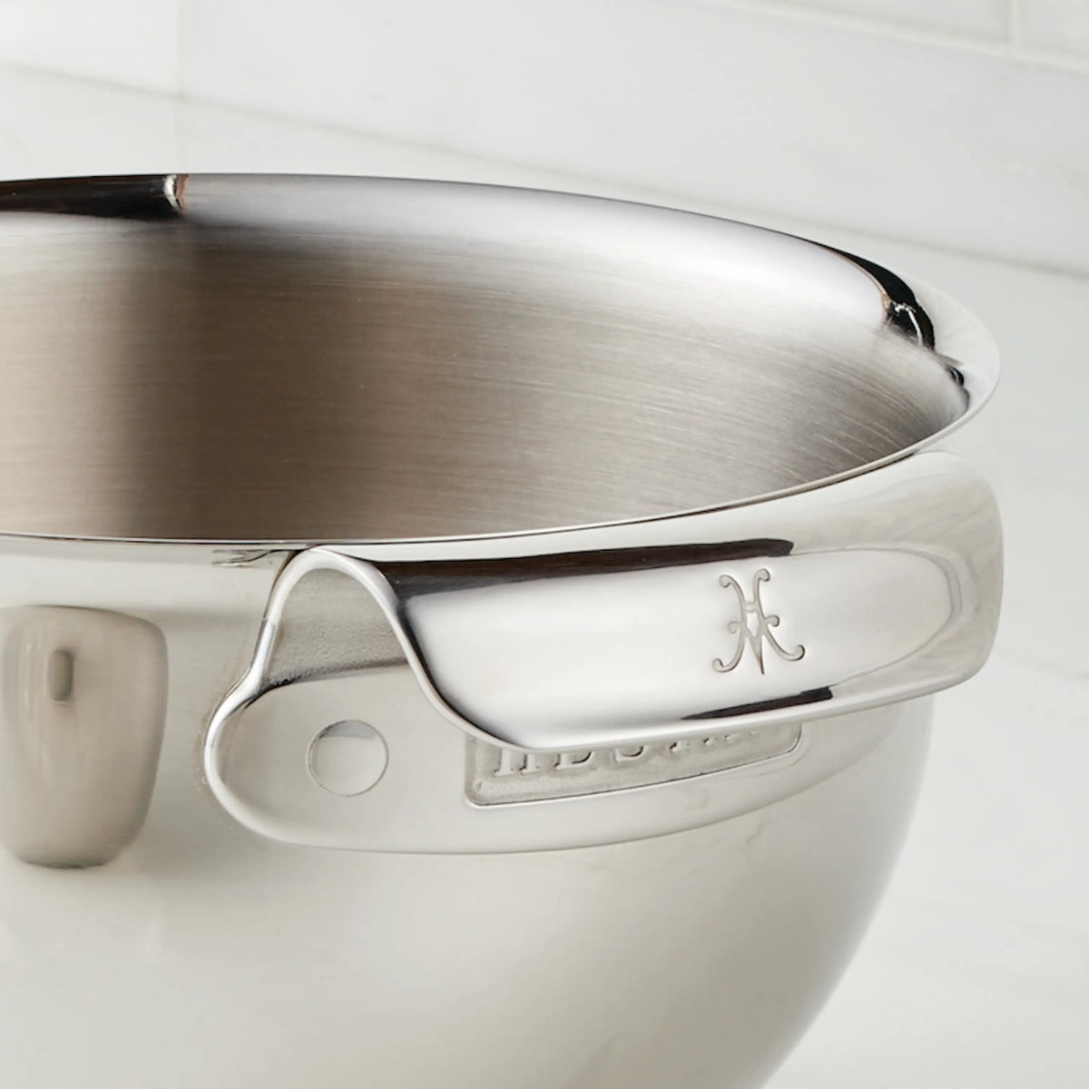 A close-up of the Hestan Provisions 7-Quart Stainless Steel Mixing Bowl with an ergonomic handle, engraved “H”, and a smooth, reflective surface. White tile background.