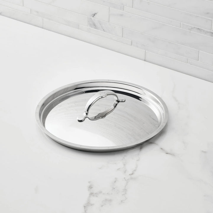 A Hestan Provisions Stainless Steel Lid with a handle rests on a white marble countertop in front of a light gray tiled backsplash.