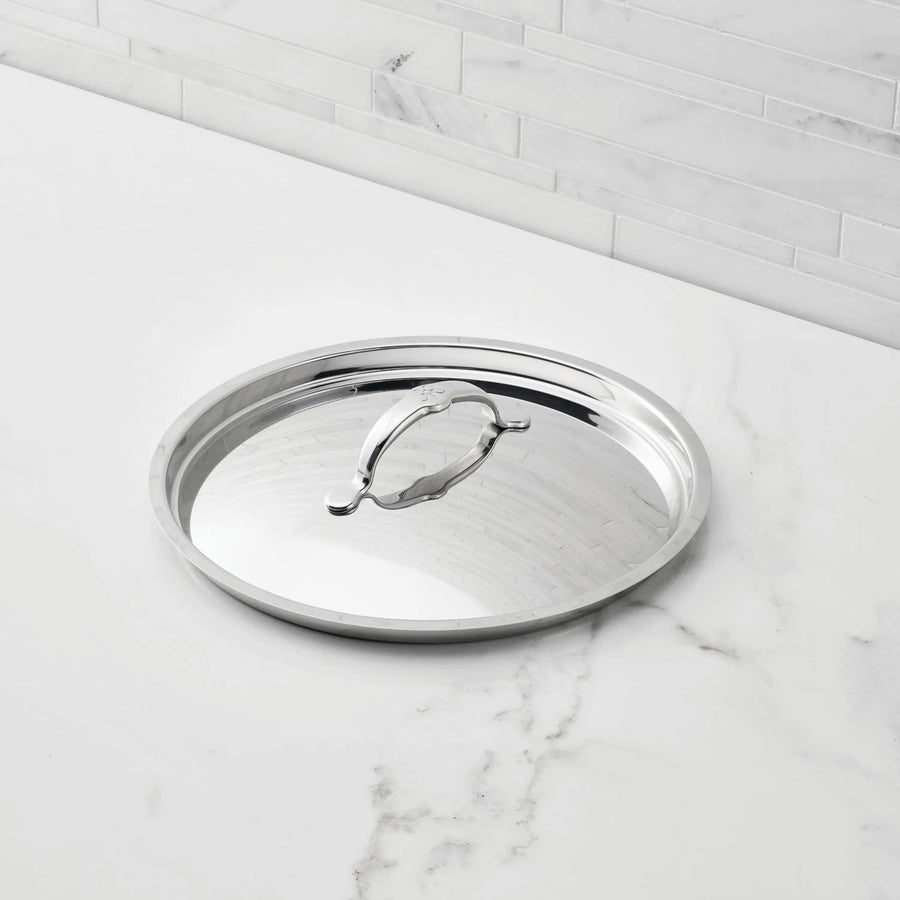 A Hestan Provisions Stainless Steel Lid with a handle rests on a white marble countertop in front of a light gray tiled backsplash.