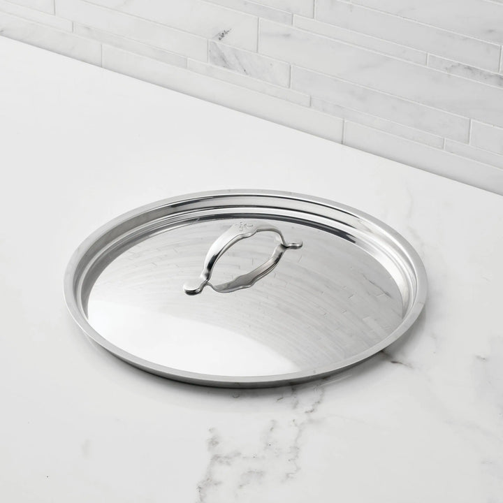 A Hestan Provisions Stainless Steel Lid with a handle and tri-ply construction rests on a white marble countertop before a light gray tiled backsplash.