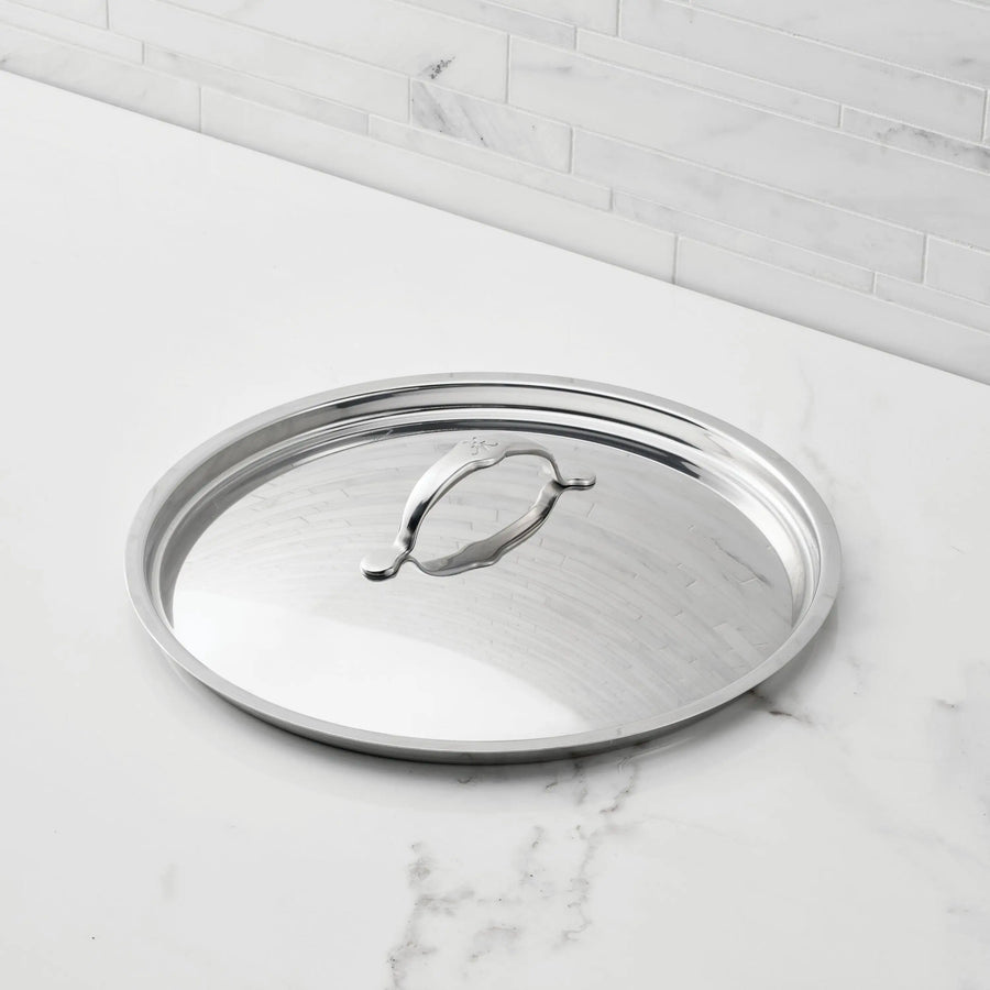 A Hestan Provisions Stainless Steel Lid with a handle and tri-ply construction rests on a white marble countertop before a light gray tiled backsplash.