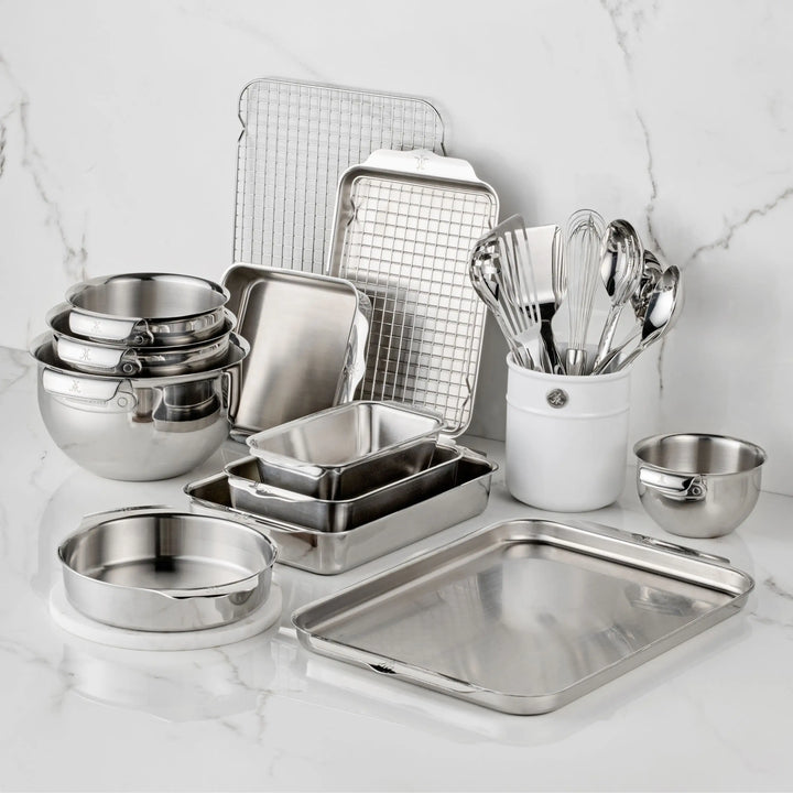 The Hestan Provisions OvenBond Tri-ply Epicurean Stainless Steel 21-Piece Set, featuring bakeware, mixing bowls, pans, a cooling rack, measuring cups, and utensils in a ceramic holder, is arranged on a white marble countertop.