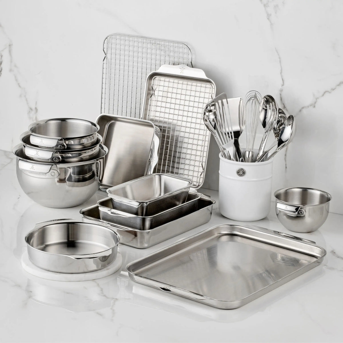 The Hestan Provisions OvenBond Tri-ply Epicurean Stainless Steel 21-Piece Set, featuring bakeware, mixing bowls, pans, a cooling rack, measuring cups, and utensils in a ceramic holder, is arranged on a white marble countertop.