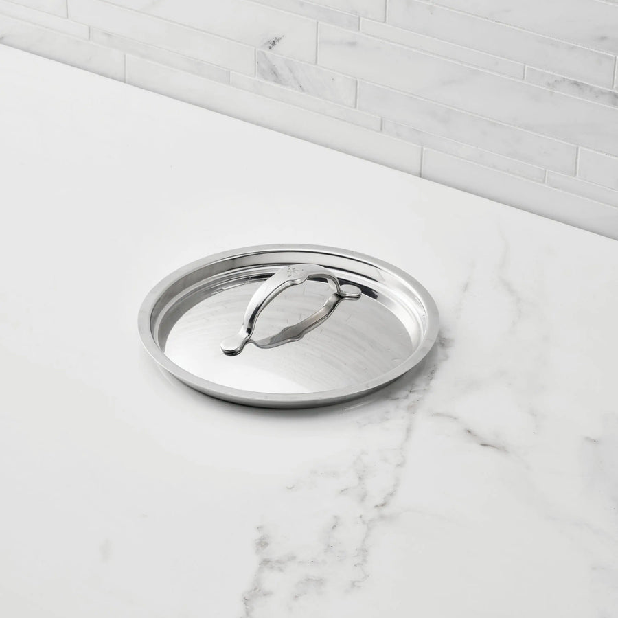 A Hestan Provisions Stainless Steel Lid with handle is placed upside down on a white marble countertop, with a tiled backsplash in the background.