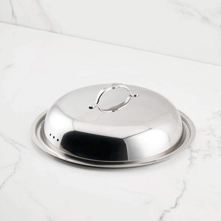 The Hestan Provisions 14" Stainless Steel Dome Lid, featuring a handle on top and tri-ply construction, sits elegantly on a matching tray atop a white marble surface.