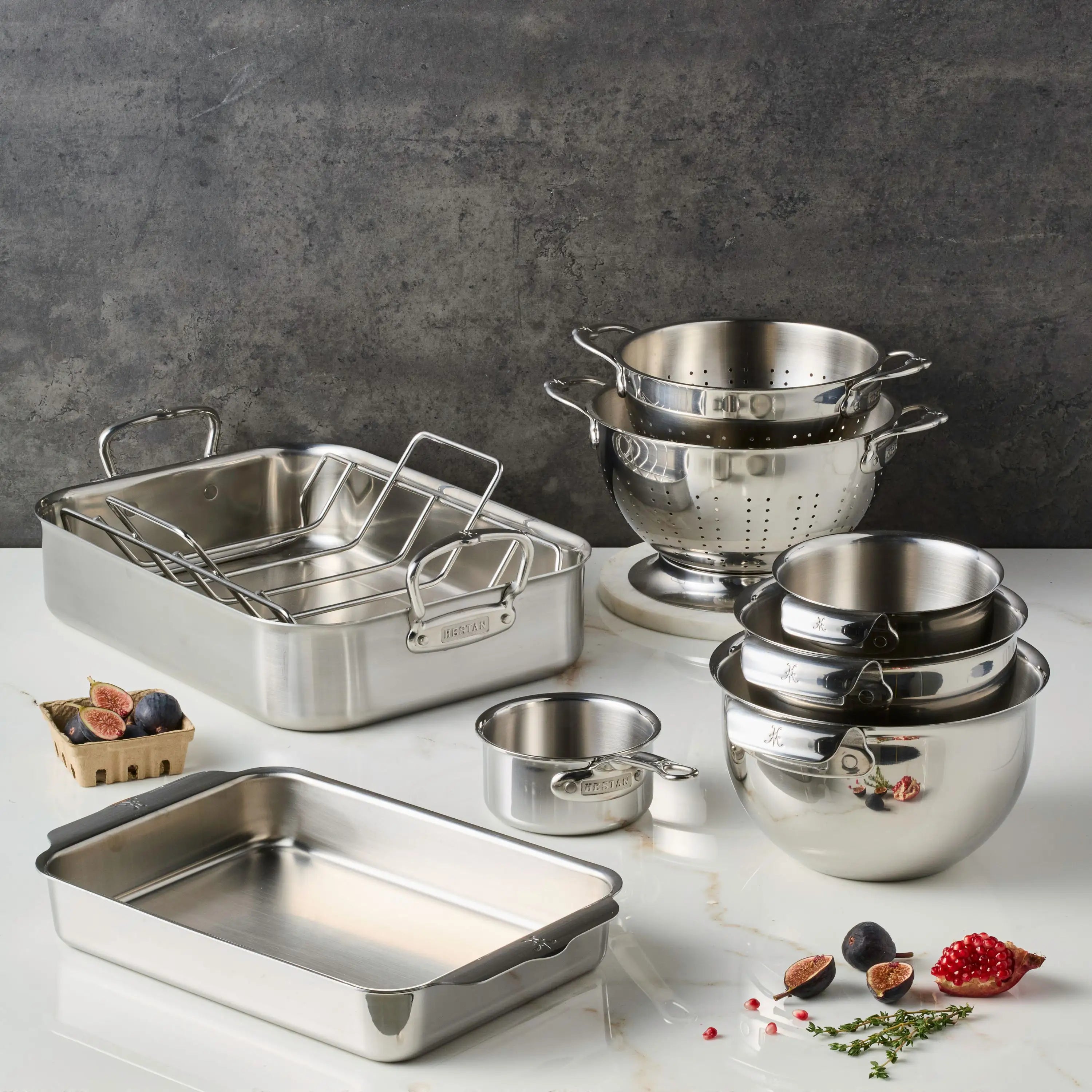 The Hestan Provisions Holiday Host and Roast 8-piece set, featuring stainless steel bakeware like mixing bowls, measuring cup, colanders, roasting pans with racks, and a baking dish—ideal for holiday prep—is arranged on a white countertop.