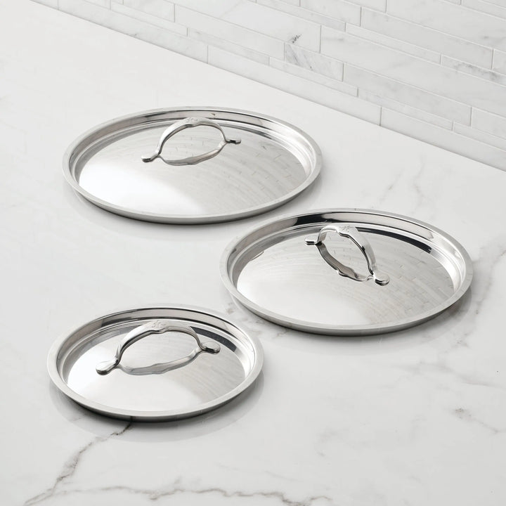 Three Hestan Provisions Stainless Steel Lids in varying sizes rest on a white marble counter with tile backsplash, each oven-safe lid featuring a central sturdy handle for both style and function.