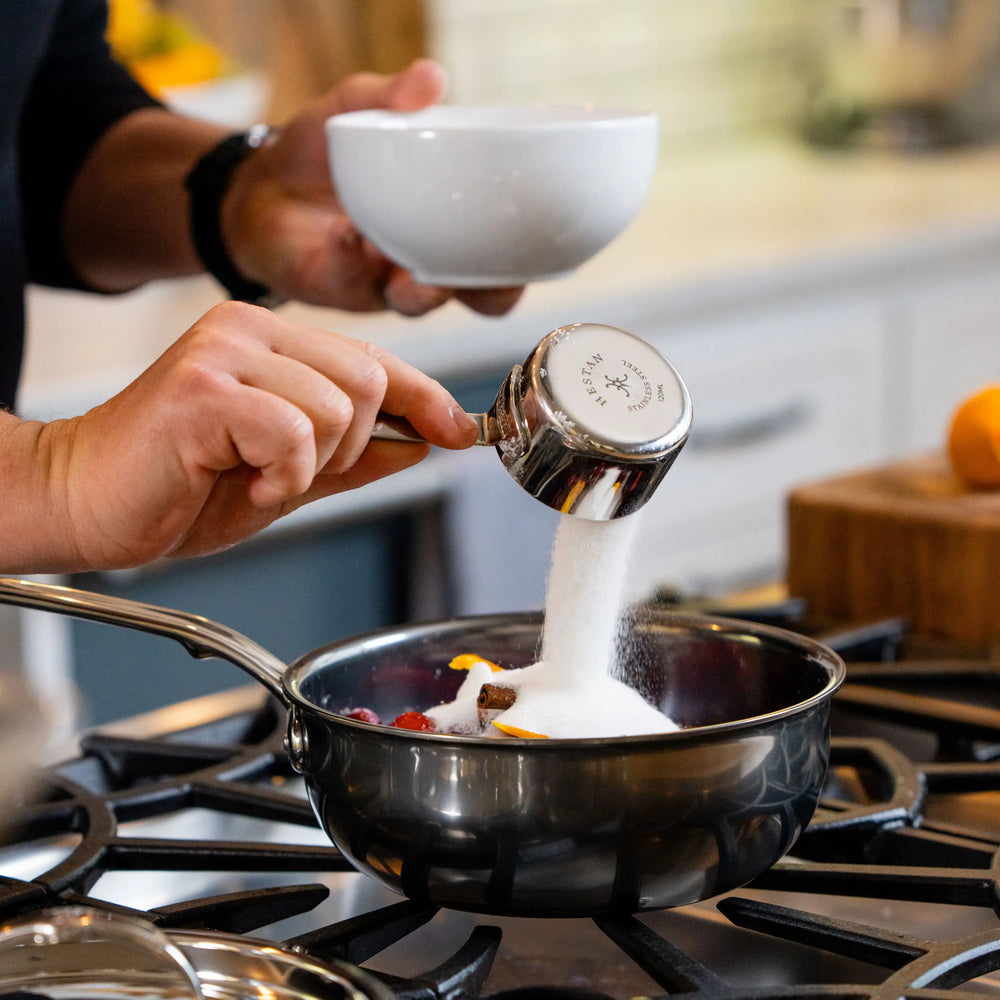 A hand pours white sugar from a Hestan Provisions® Measuring Cups and Spoons Set stainless steel measuring cup into a saucepan containing berries and cinnamon sticks on a stovetop.
