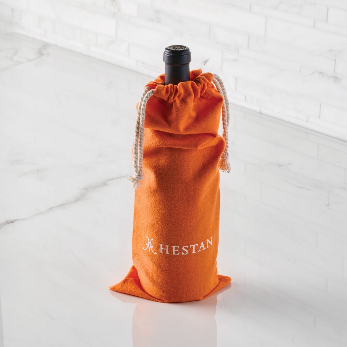 A wine bottle in an orange Accessories Flannel Cookware Storage Bag with HESTAN in white, on a white marble counter and tiled wall. Perfect for stylish cookware storage.