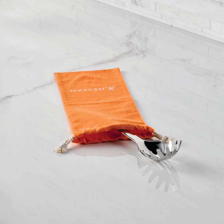 A metal pasta spoon is partly inside an Accessories Flannel Cookware Storage Bag with white drawstrings and Protective Cookware text, set on a white marble countertop.