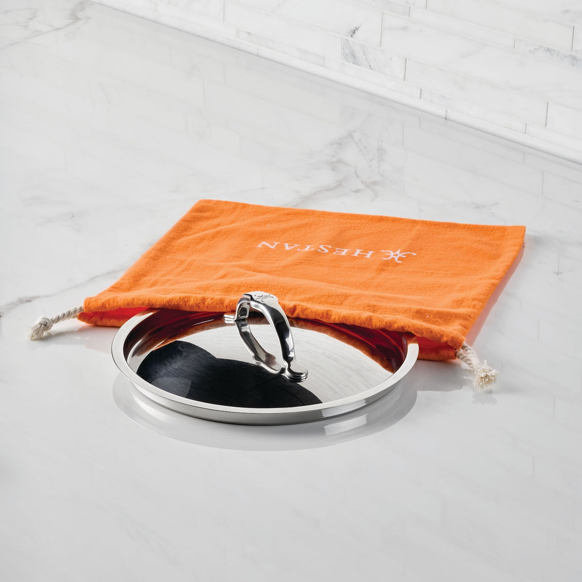 A stainless steel pot lid with a handle sits partly inside an orange Accessories Flannel Lid Storage Bag, labeled Accessories, on a white marble countertop with matching backsplash for extra cookware protection.